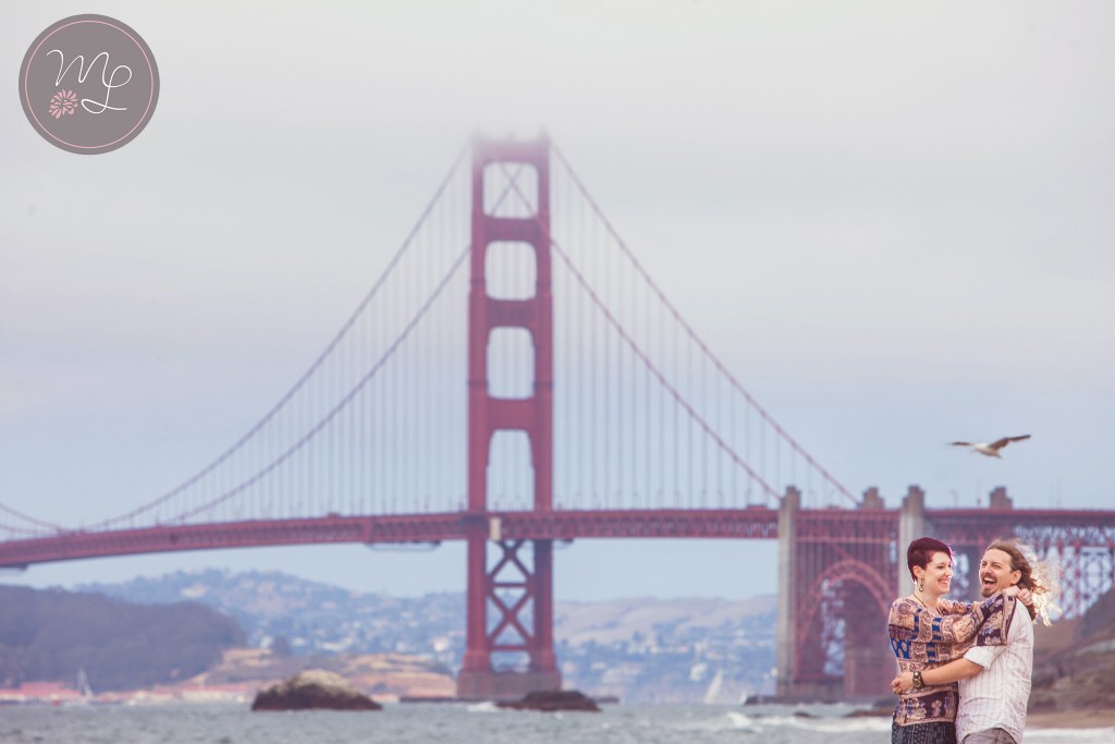 A cute couple snuggles on Baker Beach with the golden gate bridge in the background. Photography by Mabyn Ludke