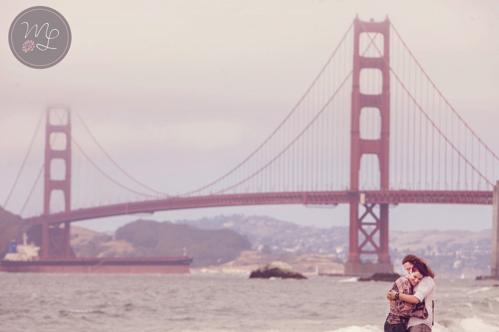 A cute couple snuggles on Baker Beach with the golden gate bridge in the background. Photography by Mabyn Ludke