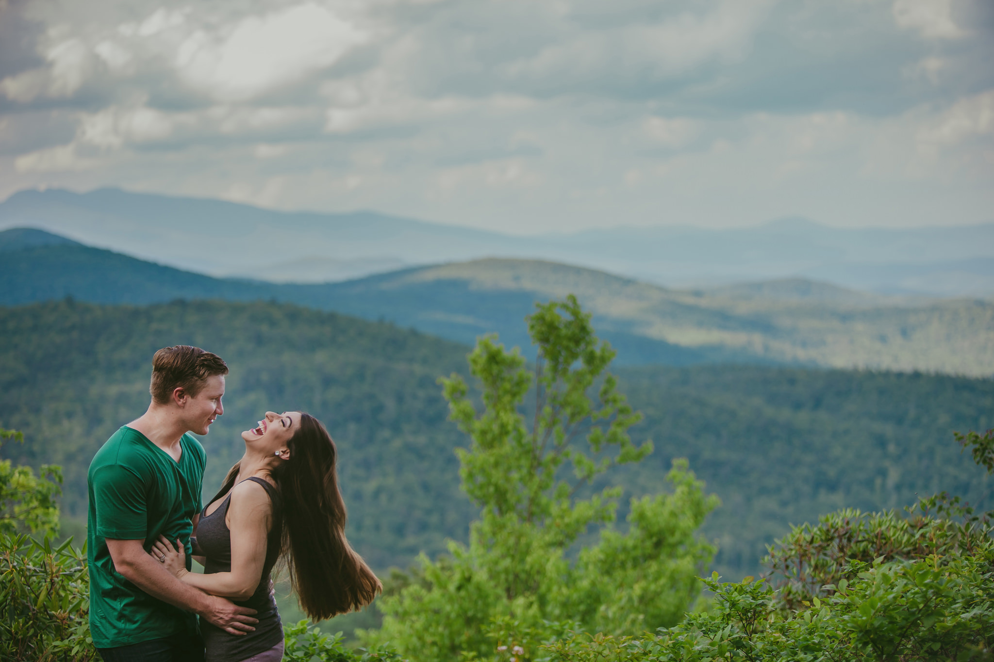 Mabyn Ludke Photography captured a beautiful couple on the Hawksbill Mountain Trail in Lineville, NC laughing with a beautiful mountin view behind them. 