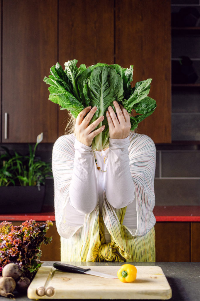 A woman wearing a white shirt covers her face with vibrant vegetables, showcasing a fun take on healthy eating.