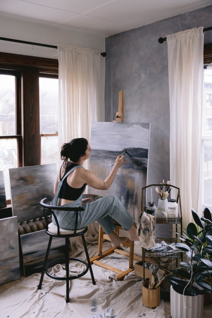 Woman sitting at an easel painting a picture while surrounded by her art and plants.