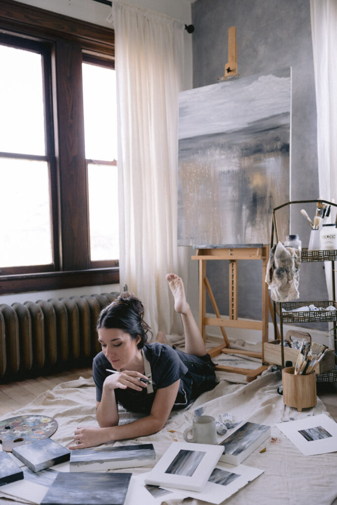 Artist Emily O'Rielly lays on the floor working on paintings surrounded by her work.