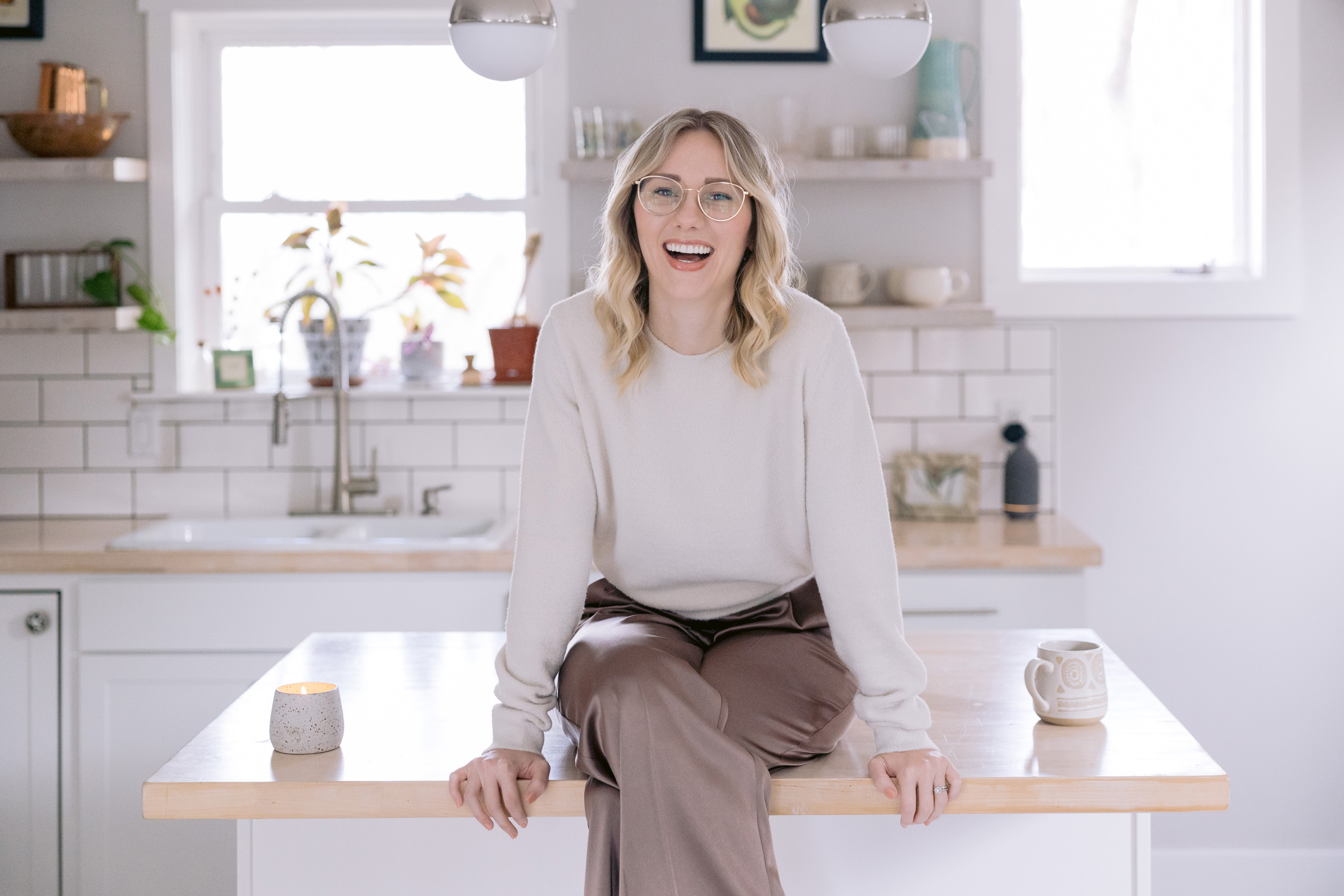 A joyful woman sits on a kitchen counter, embodying warmth and positivity in her personal brand photos by Mabyn Ludke Photography.