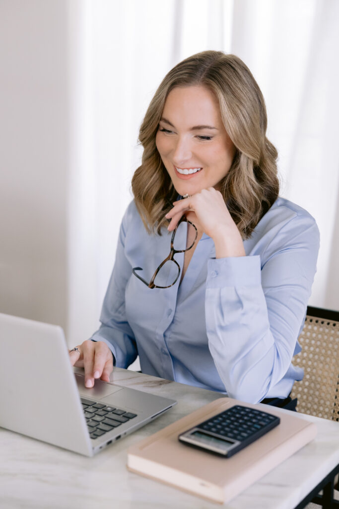 A woman in a blue shirt and glasses works at her desk with a laptop during a personal brand photoshoot.