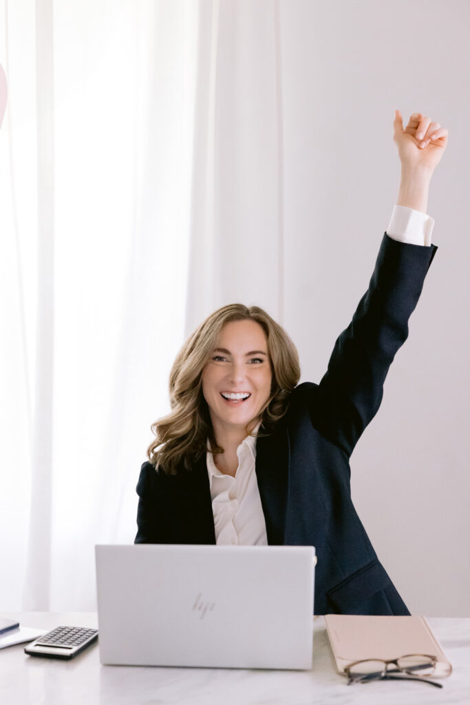 A businesswoman in a suit sits at her desk with arms raised, exuding confidence in her personal brand photoshoot.