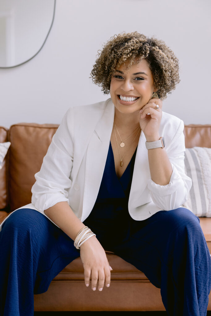 A woman wearing a white blazer and blue pants is seated on a couch, exemplifying stylish attire for professional headshots. What to wear for headshots in Charlotte NC