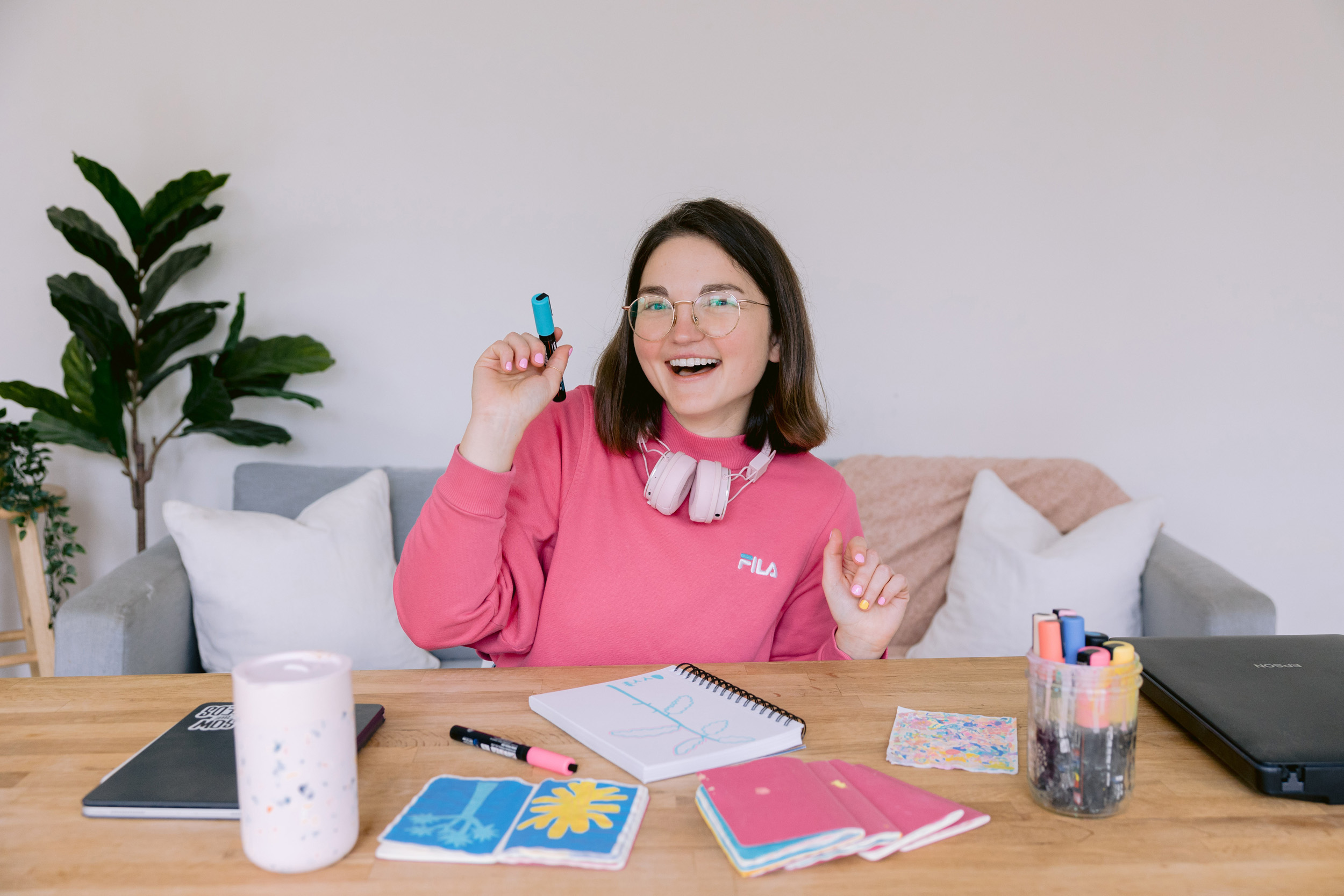 A woman wearing a pink sweatshirt is seated at a table with a notebook and art supplies, photographed by Mabyn Ludke Photography.