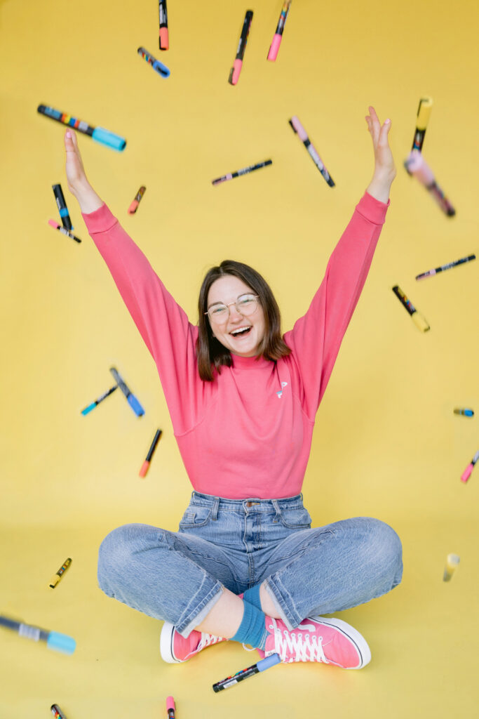 A young woman wearing a bright pink sweatshirt and jeans sits cross-legged on a yellow backdrop, smiling with her arms raised as colorful markers fall around her. Photo by Mabyn Ludke Photography.