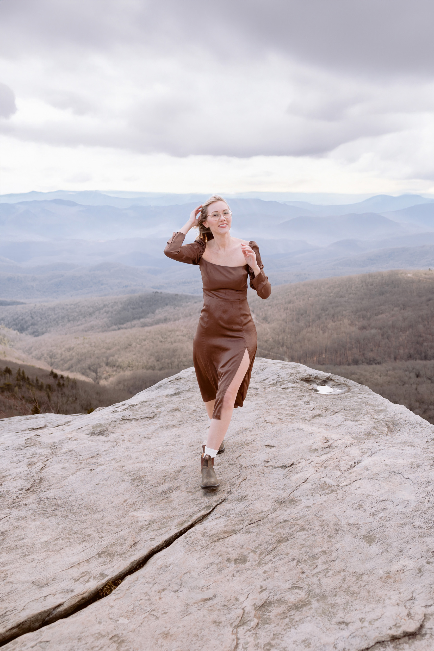 A woman wearing a brown dress poses on a rock in front of the Blue Ridge Mountains, showcasing her personal brand in a photograph by Mabyn Ludke.
