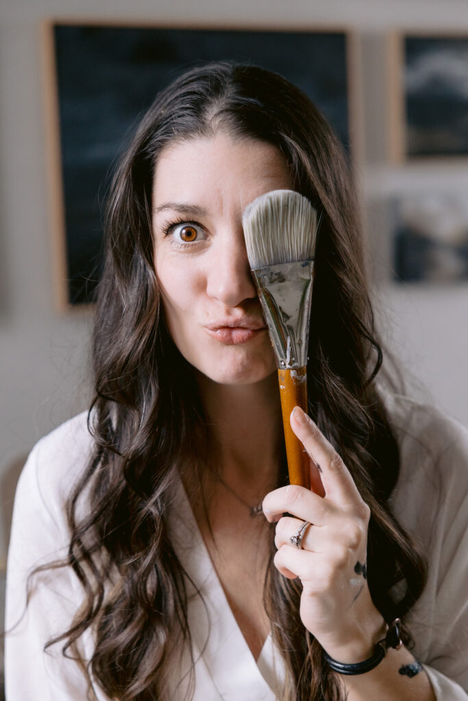 A woman poses with a paintbrush in front of her face, showcasing her artistic flair in a session by Mabyn Ludke Photography.