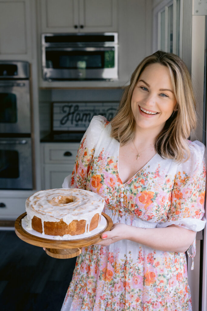 A woman wearing a floral dress stands near a window, holding a glazed bundt-style pound cake on a wooden cake stand and smiling warmly at the camera in a bright kitchen. Photo by Mabyn Ludke Photography.