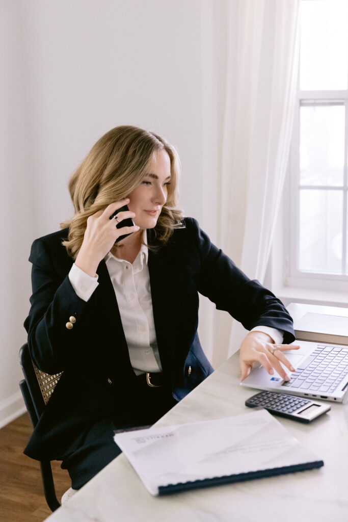A woman in a dark blazer and white blouse talks on the phone while working at a marble desk with a laptop, documents, and calculator, seated near a sunlit window with sheer white curtains. Photo by Mabyn Ludke Photography.