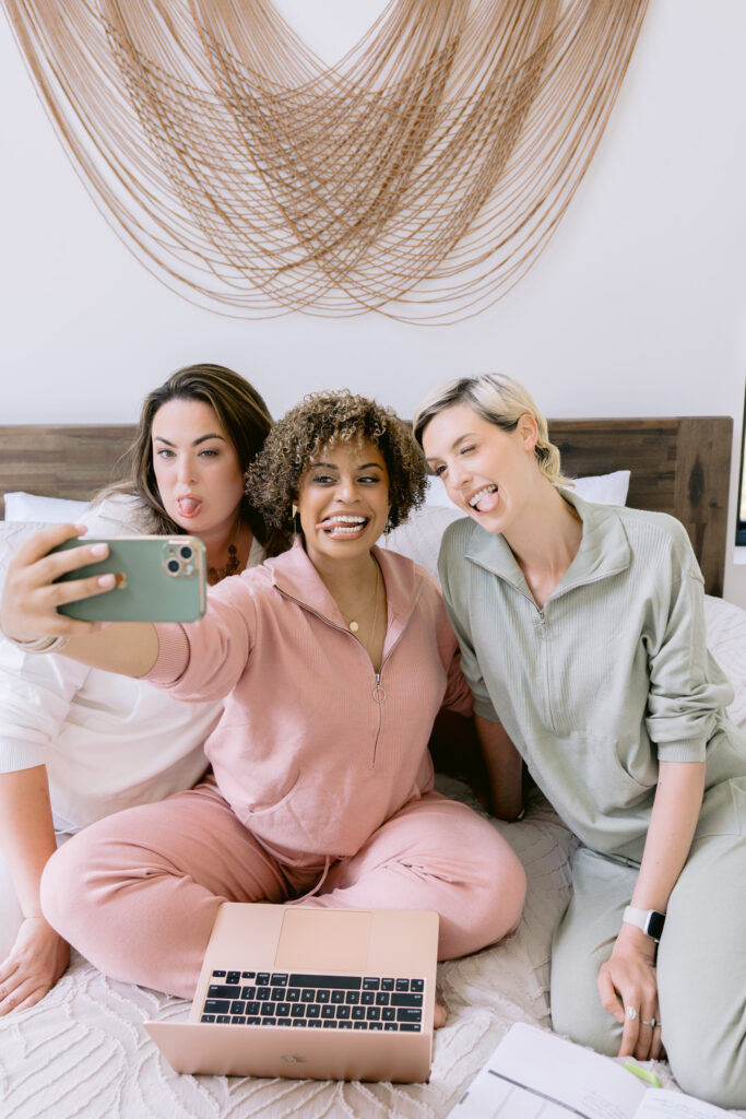 Three women in cozy loungewear sit on a bed, smiling and posing for a selfie. One holds a phone while the others lean in, with a laptop and open notebook in front of them. A woven wall hanging decorates the white wall behind them. Photo by Mabyn Ludke Photography.