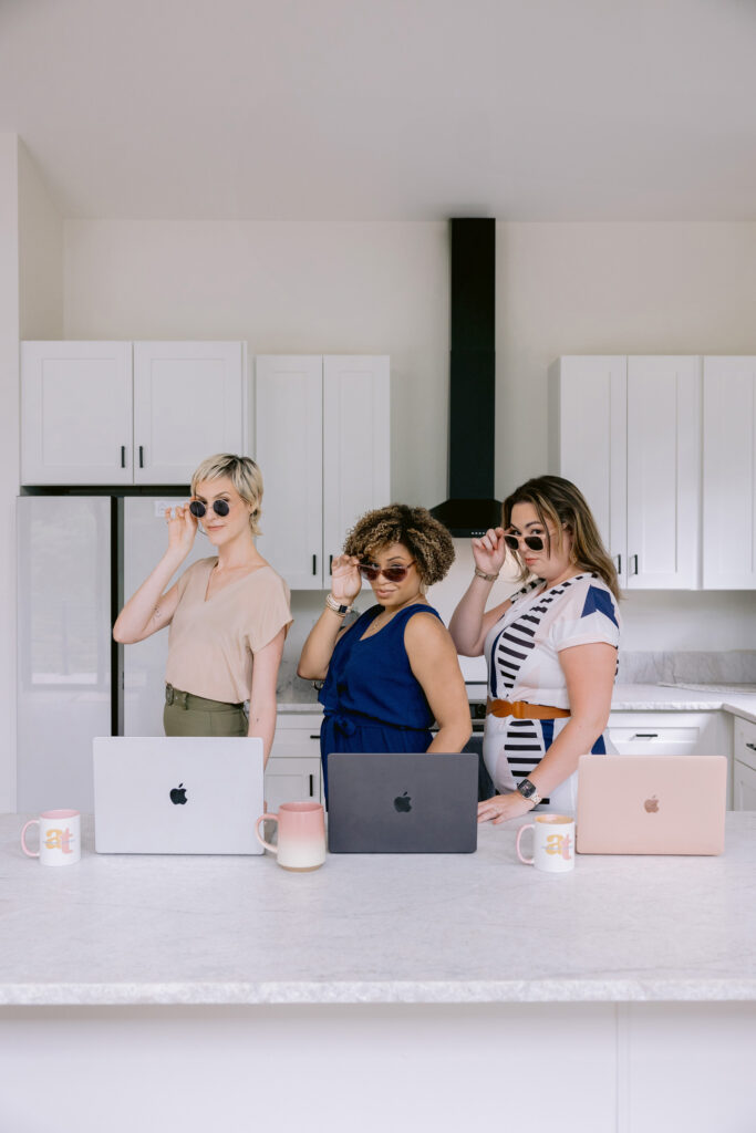Three women stand behind a kitchen island, each with a laptop and mug in front of them. They wear sunglasses and strike playful poses in a bright, modern kitchen with white cabinets and a black range hood. Photo by Mabyn Ludke Photography.
