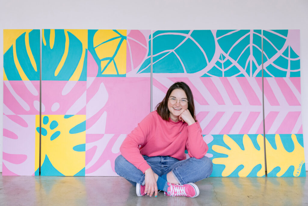 A woman wearing a pink sweatshirt and pink sneakers sits cross-legged on the floor, smiling in front of a colorful, geometric mural with bold pink, yellow, teal, and lavender leaf designs. Photo by Mabyn Ludke Photography.
