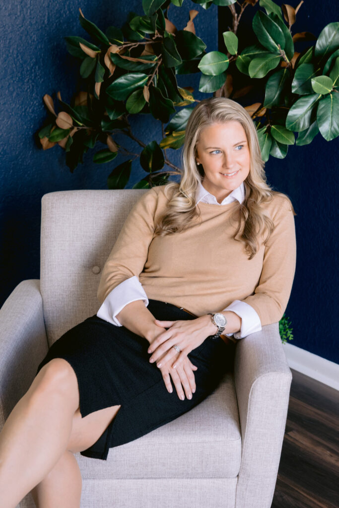 Professional woman sitting in a modern beige chair against a navy wall, posing confidently for Lake Norman brand photography.